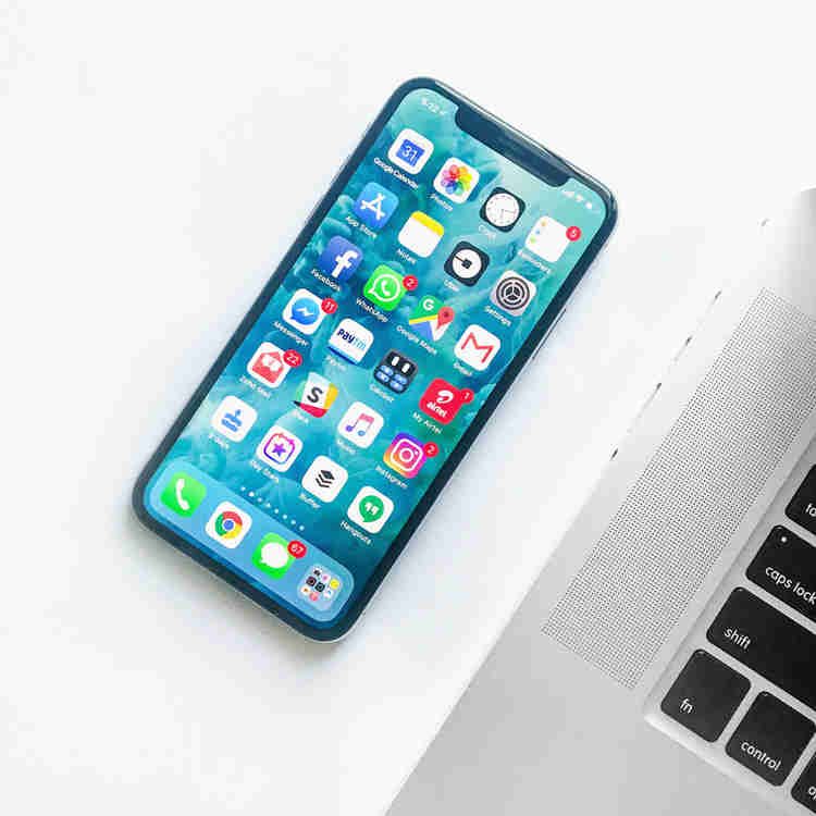 A phone sitting beside a laptop. The phone screen is on, displaying an array of apps.